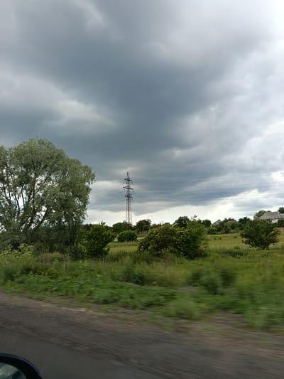 The image depicts a cloudy sky over a rural landscape. A large, leafy tree is visible on the left, and a power line tower stands in the middle ground, with power lines stretching horizontally across the frame. Several houses are scattered across the horizon, nestled among trees and green fields. The foreground shows a blurred path or road, indicating movement, and a portion of a vehicle's side mirror is visible in the lower left corner. The overall impression is of a pastoral setting under overcast conditions, possibly in or near the village of Biloziria, Ukraine.