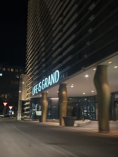 Captured at night, the image presents the "Life is Grand" building in Glattbrugg, Switzerland. The illuminated signage of "Life is Grand" is a focal point, mounted on the building's exterior. Architectural elements, including horizontal linear structures, frame the view. Nearby signage suggests a roadway.