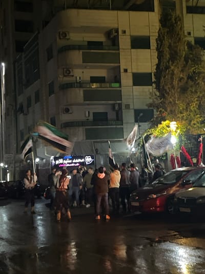 A nighttime scene depicts a gathering of people on a wet street. They hold flags, some in motion, suggestive of a protest or demonstration. A multi-story building is visible on the left, along with a red car on the right. Streetlights illuminate the scene. The presence of masks on some individuals suggests a health concern or safety measure.