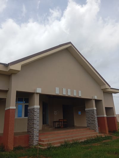 The media file shows the exterior of a light brown, single-story building with a prominent gable roof. The building has several features, including a covered entrance with a set of stairs leading up to it. The entrance area is supported by columns, some of which are decorated with grey and blue patterned tiles. To the left of the entrance, a window with blue-tinted glass is visible. The lower parts of the building's exterior walls are painted a reddish-brown color.
Inside the covered entrance, there are two wooden chairs and a small table, suggesting a waiting area or common space. On the wall of the building above the entrance, there are five rectangular white decorative elements. The sky in the background is cloudy, indicating an overcast day. The ground in front of the building is covered with green grass and some sparse vegetation. The overall scene depicts an outdoor view of a building in what appears to be a residential or institutional setting, possibly in Gwafan, Nigeria, given the provided location context. No people are visible in the frame. The time of day is indeterminate due to the overcast conditions, but it is likely daytime. There is no discernible text in the image.