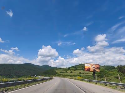 The media file depicts a scenic highway view in Potop, Bulgaria, under a bright blue sky dotted with fluffy white clouds. The time of day appears to be midday, with clear visibility. The setting is a rural, mountainous landscape, with lush green hills covered in dense forests on either side of the road. A winding asphalt road with a metal guardrail runs through the center of the frame, leading into the distance.
On the right side of the highway, a large billboard stands tall. The billboard displays an advertisement featuring a family posing with what appears to be a construction vehicle or tractor in the background. The text on the billboard is "CHIPOUND," and below it, smaller text is visible but illegible.
In the distance, several trucks are visible on the highway, suggesting ongoing traffic. The overall scene evokes a sense of travel and open road, with the natural beauty of the Bulgarian countryside providing a picturesque backdrop. There are no discernible emotions conveyed, as the media focuses on capturing the landscape and the advertisement. The weather is sunny and pleasant, indicative of a clear day. Power lines and transmission towers are also visible on the hills, hinting at the presence of infrastructure in the otherwise natural environment.