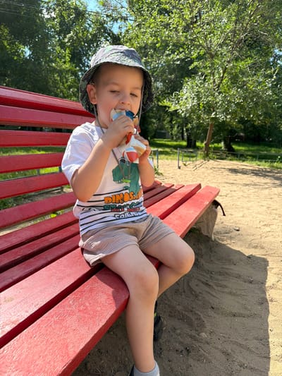 A young boy sits on a red wooden bench in a sandy park area, eating from a pouch. He is wearing a white t-shirt with a dinosaur graphic, light brown shorts, a patterned bucket hat, and grey socks. The bench is positioned on a sandy surface, with trees and green foliage visible in the background, suggesting an outdoor recreational space. The scene is illuminated by natural sunlight, creating shadows on the sand. The overall impression is a casual outdoor moment captured on a sunny day.