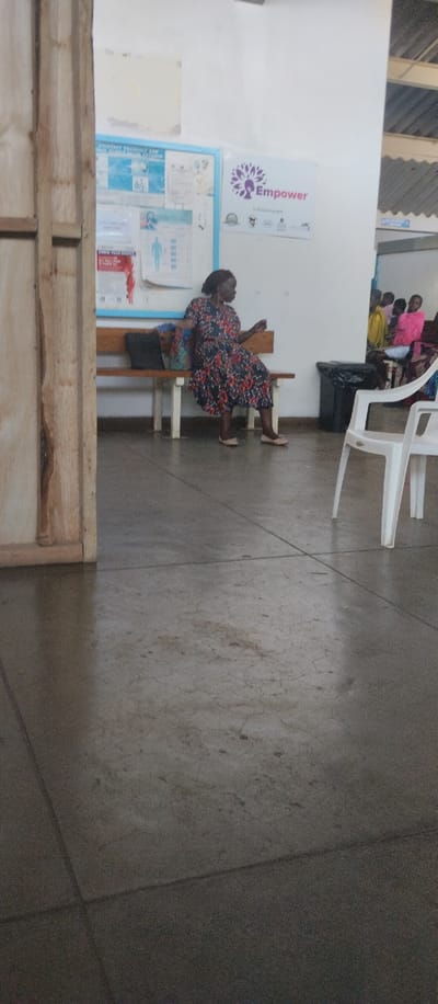The image shows a waiting area in a building located at the coordinates -0.09 latitude and 34.771 longitude in Kisumu, Kenya. A woman is seated on a wooden bench facing away from the camera. The bench is situated in front of a white wall. The wall has a bulletin board with several posters and a sign that reads "Empower." The floor is made of concrete. Two white plastic chairs are visible on the right side of the image, and a black trash can is located behind the chairs. The woman is wearing a dark dress with floral patterns. There are multiple other people visible in the background.  They are situated further away and are out of focus.  Their appearance and actions cannot be discerned.  The environment is a waiting area of a building, and the subjects are waiting to be seen or processed.