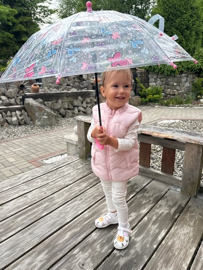 A joyful young child stands smiling brightly towards the viewer in an outdoor setting, likely a garden or patio in Wisła, Poland. The child, with light hair, wears a light pink puffer vest over a white long-sleeved shirt and white pants with subtle patterns. White sandals featuring cartoon animal faces, possibly tigers or lion cubs, are visible on their feet.
The child holds a clear plastic umbrella adorned with colorful pink and blue butterfly designs and small white speckles. The setting includes a prominent gray wooden deck or walkway where the child stands. In the background, a stone wall, lush green trees, and bushes define a landscaped area. A paved path and gravel are visible beyond the wooden structure, along with parts of a rustic wooden bench. The lighting is bright and soft, suggesting daytime. The child's broad smile and the playful umbrella convey a happy and lighthearted moment. No discernible text is visible in the image.