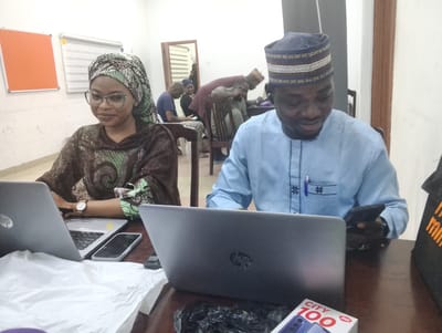 The image depicts an indoor scene in a communal workspace or office located in Giginyu, Nigeria. Two individuals occupy the foreground at a dark brown wooden table, both engaged with electronic devices, while several other people are visible in the less-focused midground and background.
On the left, a female individual with dark skin wears a patterned headwrap in green, white, and black, along with a dark brown and green patterned long-sleeved top. She wears light-framed glasses and a white-faced watch with a brown strap on her left wrist. Her gaze is directed slightly to her right, and she exhibits a subtle smile. An open silver HP laptop is positioned directly in front of her, and a silver smartphone lies face up to the right of her laptop on the table. A crumpled white plastic bag or sheet of paper rests in front of her laptop.
On the right, a male individual with dark skin wears a light blue long-sleeved traditional garment featuring dark embroidered star-like patterns on the chest and vertical lines near the placket. A blue pen is clipped into his left breast pocket. He wears a dark blue cap adorned with white and gold/tan patterned bands. He is looking down, seemingly at the dark smartphone he holds in his right hand. An open silver HP laptop is positioned in front of him. A black plastic bag is situated on the table between his laptop and the edge of the table. In the bottom right corner, a white and red product box labeled "CITY 100 itel" with an image of a smartphone is visible. To its right, a black bag with partially visible orange text is present.
The background features white walls. On the left wall, an orange rectangular board with a white frame is mounted, alongside a white horizontal window blind. In the midground, several dark wooden chairs are visible, occupied by other individuals. A person in a blue shirt is seated further left in the background, looking down at a device. Behind the foreground female, another person is partially obscured. Behind the foreground male, a person in a maroon shirt leans over a seated individual, appearing to interact with them or observe a shared item. A doorway with a brown frame leads to another white-walled room where a person is standing. The overall lighting is consistent with indoor artificial illumination.