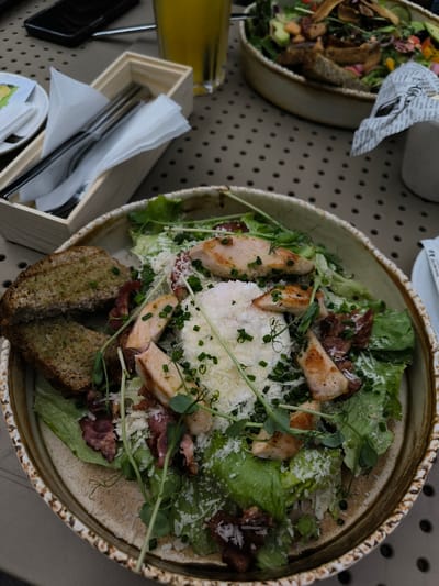 The image presents a dining table scene in Oława, Poland, viewed from a slightly elevated perspective. The primary subject is a large, shallow ceramic bowl centrally positioned, containing a Caesar salad. The salad consists of green romaine lettuce, multiple slices of grilled chicken breast, pieces of crispy bacon, and is generously topped with grated cheese and chopped chives. A poached egg forms a prominent white mound in the center of the salad. Two slices of toasted, seasoned bread are placed vertically on the left edge of the bowl. The bowl itself features a textured, light brown and cream-colored rim.
To the upper left, a light wooden caddy holds a set of silverware, including a fork, knife, and spoon, wrapped in a white paper napkin. Behind this caddy, a clear glass filled with an amber-colored liquid (likely juice) stands next to a partially visible black smartphone. A small white saucer holding a yellow packet and a folded white napkin is also present. In the blurred background on the upper right, another similar bowl, also containing a salad or comparable dish, is visible. Adjacent to it, a white condiment holder is partially obscured by crumpled paper. The table surface is dark brown with a repeating pattern of light-colored circular indentations. No individuals are visible, and no direct actions or interactions are depicted.