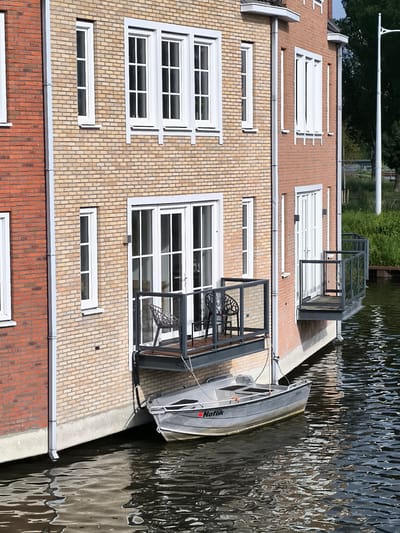 This vertical-format image presents a tranquil scene of a residential building directly bordering a canal or waterway in Alkmaar, Netherlands. The multi-story structure showcases a facade primarily composed of light beige and yellow bricks, flanked by sections of reddish-brown brick on the left and right. Numerous white-framed windows, varying in size from tall and narrow to wider with multiple panes, are set into the brickwork.
Two small, cantilevered balconies extend over the water, each featuring modern black metal railings with glass inserts. The lower-left balcony holds two black, intricately patterned outdoor chairs. Glass-paned doors provide access from the building's interior to these balconies. White drainpipes run vertically down the brick walls.
A small, light gray boat, resembling an aluminum or fiberglass dinghy, is moored directly below the left balcony. The word "Noflik" is prominently displayed in red lettering on its bow, with another partially visible word beneath it. The boat rests gently on the dark, rippling water, which reflects the daylight and parts of the building.
In the background on the far right, green trees and a slender white lamppost are visible, suggesting a green space or public area beyond the water. The scene is illuminated by bright daylight, casting clear shadows and indicating sunny or partly sunny weather. No human activity is depicted, portraying a static yet evocative picture of waterside living, characteristic of the Netherlands.