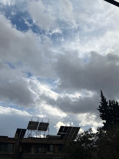 An outdoor scene in Joubar Municipality, Syria, is depicted under a sky characterized by dense, scattered grey and white clouds, with visible patches of blue sky. The lower portion of the image features the upper floors and rooftops of two adjacent multi-story buildings. On the left building's roof, two elevated metal frameworks support a total of four rectangular solar thermal collectors, arranged in two pairs. To the right, another set of dark, angled panels, likely solar collectors, are mounted directly on a lower section of an adjacent roof, alongside a cylindrical water tank. The buildings exhibit visible windows and balconies. A tall, dark evergreen tree, characterized by dense, pointed foliage, occupies the right side of the frame, partially obscuring the adjacent building's rooftop equipment. No individuals, overt actions, or direct interactions are visible within the frame. The overall light suggests daylight conditions.