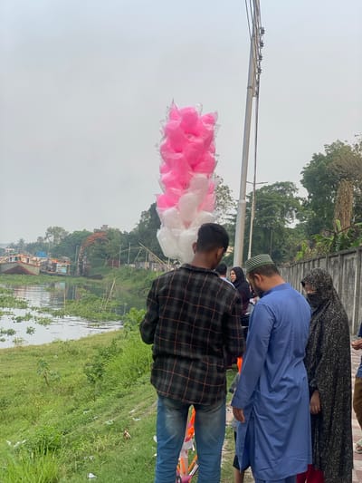 A cloudy day in Tarabo, Bangladesh, shows a row of people standing near what appears to be a river or canal. In the foreground, a man in a black and orange plaid shirt and blue jeans stands next to pink and white cotton candy. To his right, a man in a blue shirt and a small green cap stands next to a woman with a black hijab. A river runs alongside a grassy bank with green vegetation and some scattered litter. In the background, a line of buildings is seen through the vegetation. Power lines are visible against the gray sky.