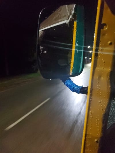 The image depicts a nighttime or low-light scene from the perspective of a vehicle, possibly in Vom, Nigeria. The foreground shows a section of a yellow vehicle body, likely a car or bus, speckled with water droplets, suggesting rain or wet conditions. A side mirror is prominently featured on the left, which is dark green with a bright horizontal yellow stripe. The arm connecting the mirror to the vehicle is covered in a textured, crinkled blue material, possibly a protective sleeve.

The reflection in the side mirror is dark and indistinct, showing what appears to be part of the vehicle's interior or another structure. Beyond the vehicle, a dark, wet road surface is visible, illuminated by what appears to be a headlight beam, creating a bright, blurred streak of light on the right side of the image, implying movement. A clear white lane marking runs diagonally across the dark asphalt in the lower right portion of the frame. There is no visible text in the image. The overall impression is of driving on a wet road during the night.