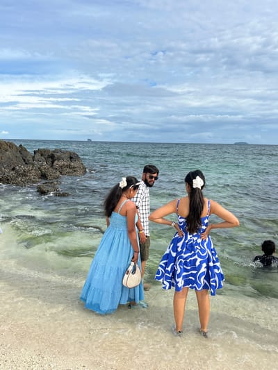 The image shows a group of people at a beach in Tambon Phru Nai, Thailand. In the foreground, two women and a man are standing in shallow water. The woman on the left is wearing a blue and white patterned dress and has her hair pulled back with a white flower. The woman on the right is wearing a blue checkered dress and is holding a light-colored handbag. The man in the middle is wearing a checkered shirt and sunglasses. In the background, a person in a black shirt is swimming in the ocean. The sky is cloudy and the water is clear with visible sand and rocks beneath the surface. The scene appears to be set during the daytime.