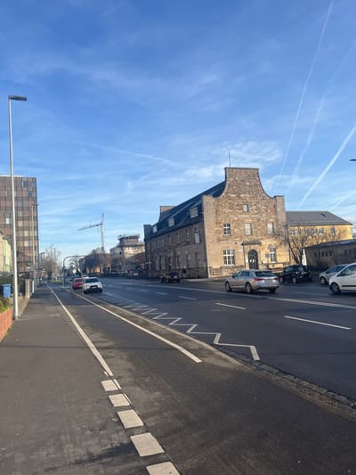 The image displays a sunlit street scene in a European city, likely Schweinfurt, Germany. The perspective is slightly elevated, looking down a wide asphalt road that recedes into the distance on the right side of the frame.

On the left, a prominent multi-story building with a distinctive stepped gable roof stands out. Its facade is constructed from light-colored stone or brick, featuring numerous rectangular windows. Beyond this, a series of other buildings, including a more modern structure with a checkered pattern, line the street. A tall construction crane is visible in the mid-distance, rising above the rooftops.

The asphalt road is well-marked with white lines. In the foreground on the right, a pedestrian crossing with broad white stripes is visible. A continuous white line borders the road, while a dashed white line and a unique zigzag white line further left indicate specific traffic or parking regulations. Several vehicles are present: a white hatchback and a black car are parked near the gabled building, and a silver car is also parked further along the street. A white van can be seen in the distance. A streetlight stands in the bottom-left foreground.

The sky, occupying the upper portion of the image, is a clear, brilliant blue with several prominent white contrails from aircraft. The bright sunlight and relatively short shadows suggest it is daytime, likely morning or early afternoon, with fair weather. Bare trees are sparsely distributed along the street, hinting at autumn or winter. No people are clearly visible, and no legible text appears on any signs, vehicles, or buildings. The scene portrays an ordinary, tranquil urban environment.