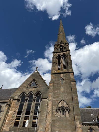 This is a low-angle shot of a church spire and part of its facade, set against a bright blue sky with fluffy white clouds. The church is constructed of weathered stone blocks, with ornate Gothic architectural features including pointed arches, tracery windows, and decorative carvings. The spire tapers to a sharp point, and the tower section has several levels of arched openings. Some windows on the main building appear to be under repair, with scaffolding and protective coverings visible. The image was taken on a sunny day.