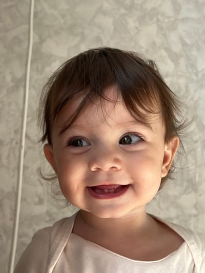 A close-up portrait shows a toddler with light brown hair and brown eyes smiling slightly with the tip of their tongue visible. The child is wearing a light pink or white shirt. The background appears to be a textured wall, possibly marble. The image seems to be taken indoors. No specific location in the city of Elena, Bulgaria can be determined from the visual information provided.