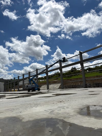 An exterior, ground-level, vertical shot captures a construction site under a bright blue sky filled with white cumulus clouds. In the foreground, a wet, light grey concrete surface reflects the sky and clouds. In the midground, a blue and grey Genie aerial work platform is positioned on the concrete. A construction worker, wearing an orange shirt and dark pants, stands in the basket of the platform, which is extended upwards towards a row of large, grey steel beams supported by vertical steel pillars. These beams form the framework of a large structure. Behind the steel framework, a lush green hillside is visible, dotted with trees and some buildings. The hillside has a retaining wall with black fencing along its top. The overall scene suggests ongoing construction work in an outdoor environment.