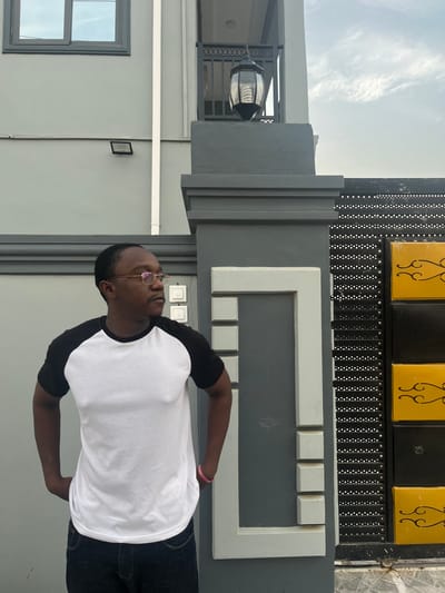 An outdoor, daytime shot shows a dark-skinned man standing in front of a modern building in Joma, Ghana. The man is wearing a white raglan t-shirt with black sleeves, dark jeans, and gold-rimmed glasses. He has a pink wristband on his right wrist and is looking upwards and slightly to the right with a neutral expression.
The background features a multi-layered architectural design. Directly behind the man is a dark gray wall adorned with a white, rectangular frame-like structure made of varying-sized blocks. Above this, a dark, perforated gate or fence panel is visible, topped by two black panels with yellow, ornate scrollwork decorations. To the man's left, a light gray wall features a dark-framed window, and a silver-colored outdoor lantern hangs from an overhang above a balcony. Another small, dark rectangular object is affixed to the light gray wall. The sky is bright with some scattered, wispy clouds, indicating clear weather. No visible text is present in the image.