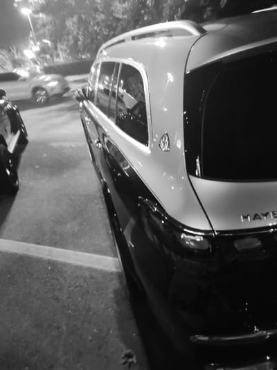 The image captures a black and silver Maybach SUV parked on a street at night. The vehicle's rear is prominently featured, with the "MAYBACH" lettering visible on the tailgate. The car is parked in a lot or on a street that appears to be in Dushanbe, Tajikistan, given the context provided. In the background, another lighter-colored SUV is visible, partially illuminated by streetlights. The environment suggests an urban setting at night, with trees and buildings faintly discernible in the background. The scene is devoid of any people.