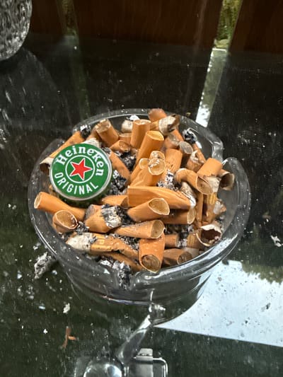 The image depicts an ashtray filled with cigarette butts, a Heineken "Original" beer bottle cap, and ash. The ashtray is made of clear glass and rests on a dark, reflective surface, possibly a table. The background shows a wooden structure and some greenery, suggesting an outdoor or semi-outdoor setting. The overall scene is one of urban detritus.
The ashtray is situated on a table in front of a window or door that looks out onto a garden area with foliage. The lighting suggests it is daytime. The ashtray is predominantly filled with stubbed-out cigarettes, many of them appearing to be filter-tipped and stained with ash. A distinctive green Heineken "Original" bottle cap, featuring the brand's red star and white lettering, is prominently placed among the cigarette butts. The glass ashtray itself has a slightly irregular, organic shape, with a rim that curves inwards. Ash and small bits of debris are scattered both within the ashtray and on the reflective surface around it.
The image is taken from a slightly elevated angle, looking down into the ashtray. The details are sharp, indicating a relatively close-up shot. There are no people visible in the image. The environment appears to be somewhat neglected or casual, given the presence of the overflowing ashtray.