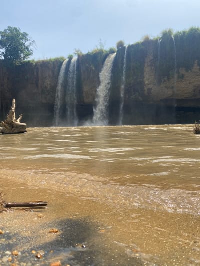 The image, when viewed in its correct orientation, captures a majestic natural landscape featuring a series of waterfalls. The foreground is dominated by a broad expanse of muddy, brown water, likely the river or stream immediately before it cascades over the cliff. This water displays visible ripples and reflections, indicating movement and possibly a sunny day. A piece of light-colored driftwood or roots is partially submerged near the upper left edge of this water body.
In the midground, multiple streams of white, foamy water plunge dramatically over a rugged, rocky cliff face. The cliff is partially covered with sparse green vegetation, including grass and small shrubs that cling to its ledges and crown its top. The turbulent waters create a misty effect at the base of the falls.
Above the cliff and waterfalls, a clear, light blue sky occupies the background, suggesting bright and sunny weather. A mature tree with dense green foliage stands prominently on the top right edge of the cliff, silhouetted against the sky. The overall bright illumination indicates it is daytime, likely mid-morning or early afternoon.
There are no people or man-made structures visible in the frame, emphasizing the pristine natural beauty of the scene. No text is discernible. This image likely depicts a scenic waterfall landmark, characteristic of the natural geography of a region like Maigizo, Nigeria, known for its diverse landscapes. The muddy color of the water suggests a river carrying a significant amount of sediment.