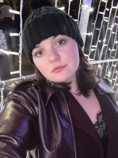A young woman with light skin and brown eyes wears a black beanie with a pom-pom, a dark brown jacket, and a lacy black top. She has a nose ring and a silver chain necklace. The background features a grid of white lights. The photo is taken at a tilted angle, with the woman looking up towards the camera.