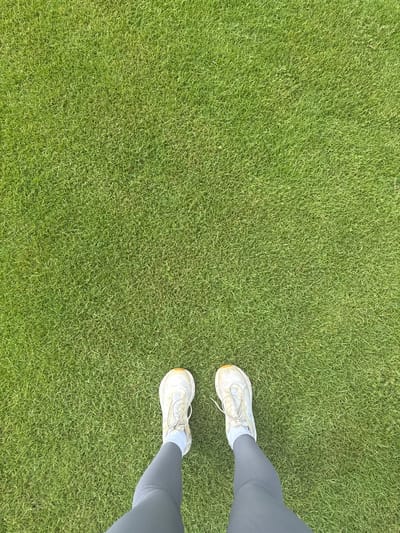 An aerial view captures a person's lower legs and feet dressed in athletic attire standing on a vibrant green lawn. The individual is wearing grey leggings and white athletic shoes with light blue socks. The lawn appears to be well-maintained and evenly cut, suggesting a park or a sports field. The scene is devoid of any urban elements, and there is no indication of the city of Miami, United States of America.