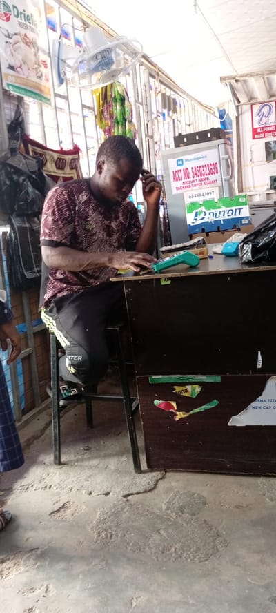 The image depicts a man sitting at a dark wooden counter in what appears to be a small shop or market stall. The man, wearing a maroon and white patterned t-shirt and black pants with yellow stripes, is looking down at the counter, with his right hand resting on his head and his left hand near a turquoise electronic device, possibly a payment terminal. The counter itself is cluttered with various items, including small boxes, a black plastic bag, and some crumpled papers. Behind the man, a white refrigerator unit is visible, displaying a "Moniepoint" label with an account number "5450323636" and an account name "BACHAFA SUPER MARKET." Above the counter, a white oscillating fan is mounted on the wall. The background shows a metal grate structure and some posters advertising products. The floor is concrete, showing signs of wear and tear. The overall setting suggests a commercial space in a potentially informal market environment in Aviation Quarters, Nigeria.