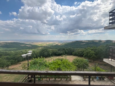 The image depicts a scenic overlook from a balcony or terrace in Pobeda, Bulgaria. In the foreground, a wooden railing frames the view. Beyond the railing, a tiered street lamp with three conical shades stands to the left. A lush green vine hangs in front of it. The middle ground showcases a verdant landscape with rolling hills covered in dense forest. A dry, grassy field with scattered trees is visible between the immediate foreground and the forested hills. In the background, a vast, open plain stretches to the horizon, dotted with what appear to be agricultural fields and distant settlements. The sky is a dynamic mix of bright blue and voluminous white clouds, suggesting a sunny day with some cloud cover. On the right side of the frame, part of a modern structure with a dark railing and a concrete balcony is visible. The balcony in the foreground is paved with hexagonal stones. A few potted plants are visible on the balcony floor.