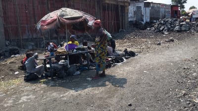 The image shows a street food stall located in Rubavu, Rwanda. The stall is set up on a gravel road in front of a building under construction,  with a pile of rocks in the background. A large umbrella provides shade for the stall. Five people are present. A woman in a colorful dress stands at the stall. Four individuals are sitting around a table that is partially covered by a red blanket. Two of the seated people are holding plates, suggesting they are eating food. The food stall is surrounded by discarded objects and waste.  The scene is captured during the day. The image has a slight blur, and the overall impression is one of informality and improvisation.
