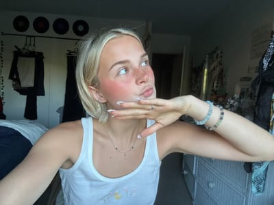 This is an undated, vertical, close-up photograph of a young Caucasian woman with blonde hair, pale skin, and freckles. She is wearing a white tank top with a small yellow duck embroidered on it, and a delicate silver necklace. Her makeup includes blush on her cheeks and lips. She has on a silver ring and several bracelets. Her fingernails are painted white with a French tip design. She is touching her face, with her eyes looking away from the camera. The background is out of focus and appears to be an indoor setting with a dresser and clothing visible. There is no discernible weather or time of day information. The location context of Whistler, Canada is provided but cannot be visually confirmed from the image itself.
