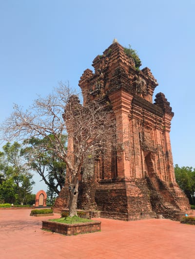 The image captures a red brick Cham tower in Tuy Hòa, Vietnam. The tower stands tall against a clear blue sky, with a bare tree in the foreground. To the right of the tower is a red brick tiled walkway and a low wall with grass. Other trees and foliage are visible in the background, partially obscuring a building with an arched opening.