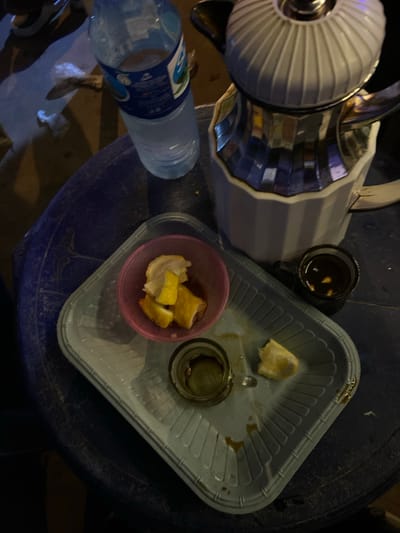 An overhead shot captures a dark, possibly outdoor, setting with a blue, circular table. On the table, a partially filled bottle of water with a blue and white label stands to the left. To its right, a silver and white coffee pot with a ridged lid sits prominently. In front of the coffee pot, a light gray plastic tray holds a small pink bowl containing pieces of lemon. To the right of the pink bowl, a small, dark-colored glass is filled with a dark liquid. Another small glass, containing a similar dark liquid and a piece of lemon, is placed further to the right on the tray. The environment appears somewhat unkempt, with visible debris and shadows indicating low lighting conditions. The location is stated to be Du, Nigeria.