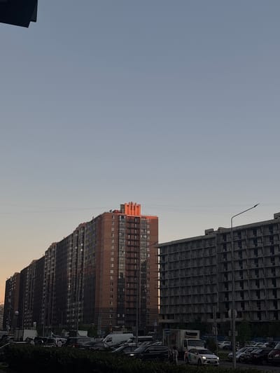 This image depicts a street scene in Murino, Russia, under a clear, pale blue sky. Two large residential buildings dominate the mid-ground. On the left is a long, multi-story brick building with numerous windows, some reflecting the sunset with an orange hue. This building is built in a terraced fashion, with different sections receding from the viewer. To the right of this building is a large, unfinished concrete structure, appearing as a skeleton of a building with many open window frames. A tall streetlamp with a curved arm is visible in front of this unfinished building.

In the foreground, a dense line of green bushes runs along the bottom edge of the frame. Behind the bushes, a street is visible, filled with a variety of vehicles. These include several cars, a white van, and a larger white truck. A few pedestrians are scattered amongst the parked and moving vehicles. The overall impression is a modern urban environment, possibly during the late afternoon or early evening, given the warm light on the brick building.