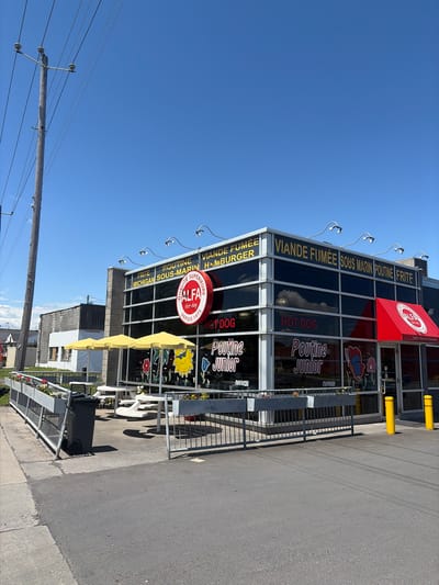 This media file captures the exterior of a fast-food restaurant, identified as "ALFA Hot-dog" at "2497 Chemin de Chambly" in Longueuil, Canada, under a clear blue sky. The multi-story building features a modern glass facade and a prominent red awning.

Several signs are visible on the building. A large horizontal sign atop the structure advertises "VIANDE FUMÉE SOUS MARIN POUTINE FRITE" (Smoked Meat Submarine Poutine Fries) and "MICHIGAN HAMBURGER". A circular red sign prominently displays "ALFA Hot-dog QUALITÉ SUPÉRIEURE DEPUIS 1971". Window decals repeat menu items like "HOT DOG" and "Poutine Junior," with some labeled "nouveau" (new).

In front of the restaurant, an outdoor seating area is visible, featuring white tables and chairs beneath bright yellow awnings. This area is separated from an asphalt surface by a black metal fence, along which planters with flowers are placed. A dark gray wheeled trash can is situated near the fence. Yellow safety bollards are positioned near the building's entrance. To the left, a wooden utility pole with numerous power lines stands against the bright, cloudless sky. The scene is illuminated by strong sunlight, indicated by the clear shadows cast by the outdoor furniture and awnings. No people are visible, and no specific event is taking place, presenting a static view of the restaurant's exterior on a sunny day.