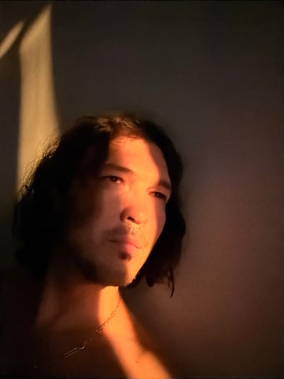 This is a close-up, waist-up, portrait photograph of a person with medium-length, dark, wavy hair. The subject is looking to their right with their head tilted slightly upwards. Dramatic lighting from the left casts a strong shadow across the left side of their face, while the right side is illuminated with a warm, orange glow, likely from natural light. The subject is wearing a thin, silver chain necklace. The background is dark and indistinct, with a vertical band of light on the far left side of the frame. There is no identifiable location in the city of Toronto, Canada.