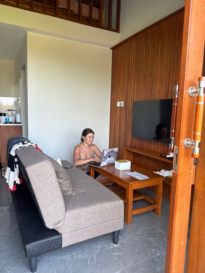 The image displays an indoor living area within a residence or accommodation located in Pecatu, Indonesia. An adult female, characterized by brown hair and wearing a leopard-print bralette or camisole, is seated on the floor to the left of a wooden coffee table. She is actively typing on an open silver laptop, which features a distinct marble-patterned skin on its lid. White wireless earbuds are visible in her ears. A smartphone rests on a gray and black two-seater sofa/futon positioned to her left, over which a striped shirt and dark garment are draped. A white square box and a piece of white paper are present on the coffee table.
The room's floor consists of dark gray tiles with a subtle marble pattern. Walls are painted light cream on the left and rear, while the right wall is covered with vertical wood paneling. A flat-screen television is mounted on the wood-paneled wall above a low wooden media console. A white power outlet with a black cable plugged in is visible on the wood wall. In the upper background, a wooden railing indicates the presence of a mezzanine level. The far left reveals a partial view of a kitchenette area, including a sink, faucet, and various containers on a counter. A partially open wooden door frame with two vertical wooden and metal handles is positioned on the far right, suggesting an entry or internal doorway. The scene is illuminated by natural light.