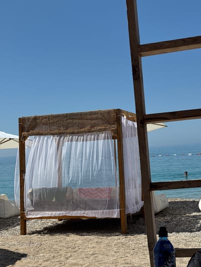 The image depicts a sunlit beach scene in Vrba, Montenegro. In the immediate foreground on the right, a weathered wooden frame structure, composed of two intersecting planks, partially obscures the view. Below this structure, a blue "Aqua Viva Sport HYDRA" water bottle with a black cap rests on the pebbled beach surface.
The central focus is a wooden beach cabana or daybed, raised on four posts. Its roof is covered with a coarse, light brown woven material. White, semi-transparent sheer curtains enclose the cabana, providing partial privacy. Inside, a bed or platform is visible, furnished with a green fabric item on the left and a red and white patterned towel or blanket on the right.
To the left of the cabana, a white beach umbrella is partially visible, deployed above the horizon. A white beanbag-style cushion is situated on the pebbled beach to the left of the cabana, and another is partially visible to the right, behind the cabana. The ground consists of a mixture of sand and small pebbles/gravel.
In the background, the calm, clear blue-green sea extends to the horizon. A line of red and white cylindrical buoys is visible across the water, marking a designated area. To the right, a person's head and shoulders are visible as they swim or stand in the sea. The sky above is clear and light blue, indicating bright, sunny weather.