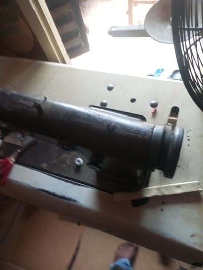 A low-angle, slightly blurry, medium shot captures an industrial setting, likely a workshop or repair shop in Agbara, Nigeria. In the foreground, a dark, cylindrical object, possibly part of a sewing machine or industrial equipment, lies horizontally across a light-colored, textured surface that appears to be a workbench or table. The object is worn and shows signs of use, with a rough, dark texture. Several small, dark circular indentations, possibly screw heads or rivets, are visible on the workbench.

To the right of the main object, a black fan with visible blades is positioned, partially cut off by the frame. Its presence suggests a need for ventilation or cooling in the workspace. In the background, the scene is dimly lit, with parts of wooden structures and a bright, possibly natural, light source suggesting an open area or a window. The overall impression is one of a functional, working environment where repairs or manufacturing might take place. A foot, clad in blue denim, is visible in the lower right corner, indicating human presence in the scene. The environment appears cluttered, with some red material visible on the left side of the main object.