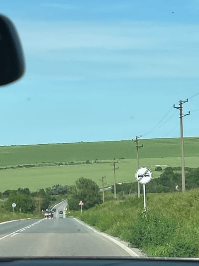 The image, taken from inside a vehicle, presents a rural road scene under a bright, clear blue sky. The perspective is rotated 90 degrees clockwise, showing the road curving gently through a countryside landscape.
On the left side of the road, extensive green agricultural fields stretch towards the horizon, interspersed with multiple wooden utility poles carrying power lines. The road itself is asphalt, marked by a solid white line separating the lanes. Ahead on the road, a line of vehicles, including what appears to be cars and possibly a truck, can be seen following the road's curve. The right side of the road is bordered by lush green vegetation.
Several European road signs are visible. A prominent circular regulatory sign features a red border with a diagonal red slash across two car silhouettes (a black car and a red car), indicating "No Overtaking" for all motor vehicles. Further down the road, a triangular warning sign with a red border and a black exclamation mark signifies "Other Dangers." Another circular sign, likely marking the end of a prohibition, is visible in the distance.
The scene depicts bright daytime conditions with clear weather. No people are explicitly visible. A dark object, possibly part of the vehicle's interior or a reflection, appears in the bottom left corner. The setting is an open countryside road, specified to be near Kumanovo, Bulgaria.