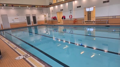 The image shows an indoor swimming pool, likely in a public facility such as a community center or gym, located in Markham, Canada. The pool is divided into lanes by floating markers, suggesting it is set up for lap swimming or training. The water is a clear, bright blue and reflects the overhead lighting.

Around the pool, there are several notable objects and features. Along the walls, there are changing room doors, a clock, and signs. One sign clearly reads "DEEP AREA" above a white board with some writing on it, likely rules or information. Another sign, partially visible, says "SHALLOW WATER NO DIVING". Several life jackets in red and blue are hanging on hooks on the wall, and a red life preserver ring is mounted above a small, bright orange chair. Benches are installed along one side of the pool area. The floor around the pool is tiled with a mosaic pattern in shades of orange, yellow, and brown.

The lighting in the room appears to be artificial, coming from a series of ceiling lights that are reflected in the water. There is no indication of the time of day or weather outside. The scene appears quiet and empty, with no people visible, suggesting the pool is either closed or not in use at the moment the photo was taken. The overall impression is one of a clean, functional, and well-maintained swimming facility.