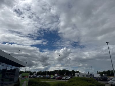 This media file depicts an outdoor scene at what appears to be a service station or rest stop. The foreground features a modern building with large glass windows, reflecting the sky. To the right of the building, a tall lamppost stands, and beyond that, a parking lot filled with various vehicles, including cars, vans, and a large truck. Greenery, consisting of bushes and trees, surrounds the parking area.

The sky is a dominant element, filled with a dramatic display of clouds. Patches of blue sky are visible between the voluminous, grey and white clouds, suggesting a changeable weather pattern. The lighting indicates it is daytime.

There are a couple of birds in flight, adding a dynamic element to the scene. One bird is visible in the upper left quadrant, and another is more centrally located in the upper half of the image.

A signpost is visible in front of the building, displaying logos for various food establishments: "Applegreen," "Bakewell," "Burger King," "Lavazza," and "Subway." This text clearly identifies the location as an Applegreen service station, likely in Ireland given the location context.

The overall impression is one of a typical day at a travel stop, with the dynamic sky providing a sense of atmosphere. There are no people clearly visible in the image.