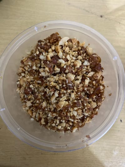 A top-down, close-up shot displays a clear, round plastic container filled with a mixture of finely chopped ingredients. The contents consist of two primary components: irregular, dark brown, somewhat translucent pieces resembling chopped dates, and lighter, off-white to light brown, angular fragments that appear to be chopped nuts (possibly peanuts, cashews, or almonds). The two ingredients are thoroughly mixed and fill the container almost to its rim, showcasing a textured, granular appearance.

The transparent plastic container has a simple design with a visible rim. It rests on a light-colored surface, which shows subtle grain and some darker specks, suggesting a wooden table or countertop. There are no people visible, and no specific activity is taking place other than the static presentation of the food item. No text is discernible on the container or in the background. The scene depicts an indoor setting, likely a kitchen or dining area, with no distinct geographical cues to confirm the Bekasi Regency, Indonesia, location beyond the generic indoor environment. The mixture appears to be a prepared ingredient or a snack component.