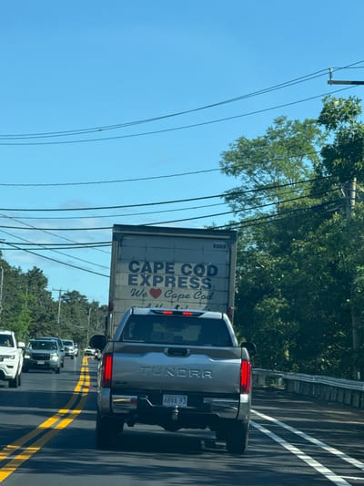 On a sunny day, a silver Toyota Tundra pickup truck is driving on a two-lane road in Wellfleet, United States of America. The truck is in the right lane, and a box truck with "CAPE COD EXPRESS" written on it is in front of the Tundra. The Tundra's license plate reads "6BVA 93". Other vehicles are visible in the background, including a white SUV in the left lane. The road is lined with trees on the right side and utility poles and power lines are visible in the sky.