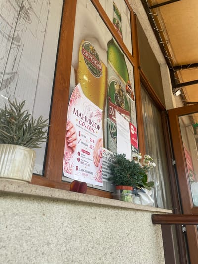 The image displays an eye-level, slightly upward-angled view of a building's exterior, focusing on a window display and adjacent architectural elements in Konstantinovo, Bulgaria.
The central feature is a window with dark brown wooden frames. The left-most pane is covered by a fine mesh screen, partially obscuring the view through it. A light-colored, ribbed ceramic pot containing a spiky green plant is positioned on the windowsill on the left side.
Several posters are affixed to the windowpanes. A large advertisement for "БУРГАСКО" (Burgasko) beer is visible, depicting a glass of beer with foam and a green beer bottle. Overlaid or adjacent to this is a pink and white poster for a "МАНИКЮР салон" (Manicure Salon). This poster illustrates manicured hands with pink and glitter nails, and a pair of feet with pedicured toenails. The text details services: "ГЕЛ ЛАК" (Gel Lacquer) for 25 € and "ИЗГРАЖДАНЕ" (Build/Extension) for 50 €. Contact information includes a Facebook page "Dream Nails by Nenka Ilieva" and a Viber/WhatsApp number: 0897 471 367. The salon's location is specified as "с. КОНСТАНТИНОВО ЦЕНТЪР" (village Konstantinovo, Center). To the right of the manicure poster, several white papers and a red card with the "Edenred" logo are partially visible, also attached to the window.
On the windowsill, between the two potted plants, two reddish-purple peaches or nectarines are resting. To the right of these fruits, another potted plant with dense green foliage and small white flowers is present in a dark pot. A crumpled white item is located behind this plant.
The wall beneath the windowsill features a light-colored, granular, textured finish, consistent with stucco or pebbledash. Above the window and extending to the right, a beige-colored ceiling or canopy structure is visible, with multiple metal hooks and electrical wiring running along its edge. A small, white, rectangular outdoor light fixture is mounted on the wall below this canopy, to the right of the window frame. An open wooden-framed door is partially visible on the far right, revealing a darker interior space.