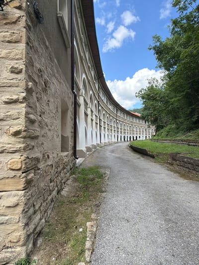 The image depicts a curved, multi-story building with a series of arches along its facade. The building is constructed with a combination of rough-hewn stone on the lower section and a lighter-colored material with decorative white trim on the upper sections. A dark gutter pipe runs vertically down the side of the building. A gravel path curves away from the building and into a densely wooded area on the right side of the frame. The sky above is blue with scattered white clouds. The scene appears to be outdoors and in a somewhat overgrown natural setting, possibly a historical site or park. This location is identified as the ruins of the National Preslav Historical-Cultural Reserve in Veliki Preslav, Bulgaria. There are no people visible in the image.