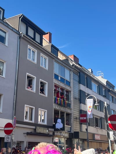 The image shows a building in the city of Köln, Germany, at the coordinates 50.926 latitude and 6.958 longitude. The building is made of gray stone with multiple windows. There is a man leaning out of one of the windows, waving. Another man is visible leaning out of a window above him. Both men are wearing shirts and appear to be watching a parade taking place in the street below. The building is in the middle of the image and the rest of the image is filled with the sky, other buildings, and a crowd of people.  A street sign with a no-entry sign and the number 39 is visible in the bottom right corner.  There are other signs, including one for a cafe and one for a grocery store. There is a sign with a logo on a street lamp. The crowd is partially visible, and some are wearing costumes. The sky is blue with some clouds.