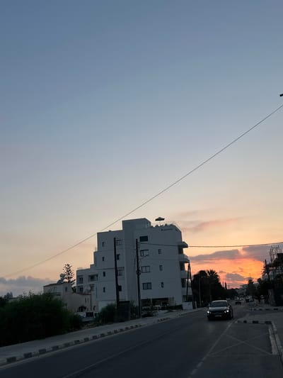 The image depicts a street scene in Paphos Municipality, Cyprus, during what appears to be sunset or dusk. A modern, white, multi-story building with balconies and a rooftop area is prominently featured on the left side of the frame. The building has the words "Island Blue" visible on its facade.
A dark-colored sedan is driving on the road in the foreground, its headlights illuminated. The road has lane markings and a sidewalk with a distinctive gray and white border. To the right of the road, there is another building under construction with scaffolding.
The sky above is a gradient of pale blue at the top, transitioning to soft pinks and oranges near the horizon, indicating sunset. Several trees and other buildings are visible in the background, suggesting an urban or suburban environment. Overhead power lines are also present, crisscrossing the sky. The overall lighting suggests the late afternoon or early evening.