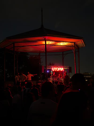 The image depicts an outdoor evening event at the Mordecai Richler gazebo in Montreal, Canada. A crowd of people, mostly silhouetted and appearing in dark clothing, are gathered in front of a stage set up inside the gazebo. The stage is illuminated by red and blue lights, with a prominent neon sign spelling out "UTOPIA" in red. Two DJs are visible behind a console, actively engaged in their performance, while other individuals stand around them. Above the stage, another neon sign spells out "MORDECAI RICHLER" in orange. The gazebo itself is red-lit from within, casting a warm glow. The background is a dark night sky with some distant, dimly lit structures visible, suggesting an urban setting. The overall atmosphere appears to be a lively outdoor concert or DJ set.