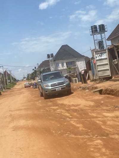 The image shows a dirt road in Iddo Tudunwada, Nigeria. A silver Toyota RAV4 is parked on the right side of the road, facing forward. Behind the RAV4, a motorcycle is parked. Further down the road, another car is visible. Buildings line the sides of the road, some with water tanks on elevated platforms. The sky is blue with scattered clouds. The road surface is predominantly red dirt, with some areas showing signs of wear and tear.
