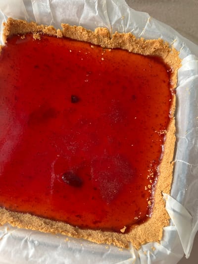 A close-up, overhead shot shows a rectangular food item within a baking dish lined with white parchment paper. The food item consists of a golden-brown, crumbly crust forming the base and sides. This crust encloses a vibrant, translucent red filling, likely a fruit jelly or glaze, with small dark specks dispersed throughout, indicating the presence of fruit pulp or seeds. There are a few slightly darker, irregular shapes within the red filling, possibly whole berries or concentrated fruit pieces. The surface of the filling is slightly uneven, with some areas appearing smoother and shinier than others, suggesting it might have been recently poured or is still setting. The lighting highlights the texture of the crust and the glossy sheen of the filling. There is no identifiable location in Antalya, Turkey, as the image solely focuses on the food item.