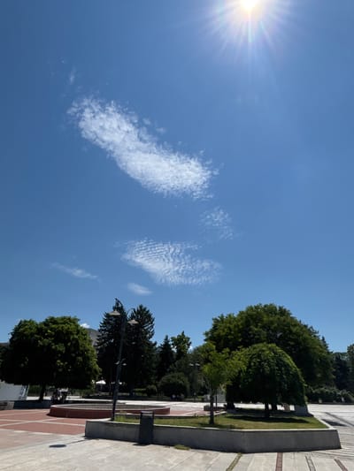 This is an outdoor, eye-level, vertical shot of a public square in Silistra, Bulgaria, on a sunny day. The sky is a vibrant blue, with a bright sun in the upper right corner casting rays. There are two distinct cloud formations in the sky; one is a long, wispy trail, and the other is a textured, patterned cloud.
In the foreground, a paved plaza with a red brick border surrounds a circular, raised concrete planter filled with grass. To the right of the planter, a concrete bench with a trash receptacle sits. A lamppost with two lights stands near the center of the image, positioned behind the planter.
The middle ground features a variety of trees, including a large, rounded tree on the left, several tall, conical evergreen trees, and a weeping willow-like tree on the right. In the background, partially obscured by trees, are buildings and what appears to be a fountain. A few individuals can be seen in the background, with one person on a bicycle near the large tree on the left. The overall scene depicts a serene urban park setting.